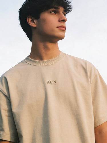 Man wearing a beige crewneck t-shirt with subtle embroidered text on the chest, photographed outdoors.
