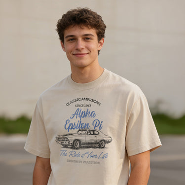 Young man wearing a vintage-style beige graphic T-shirt featuring a retro muscle car illustration and cursive text, standing outdoors with a neutral blurred background.