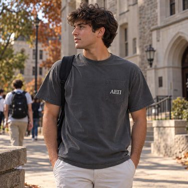 College-aged male wearing a charcoal Comfort Colors pocket tee with white heat-sealed Greek lettering on the front pocket, photographed on a college campus.