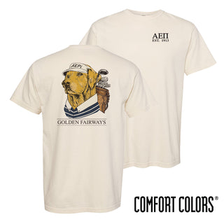 Lifestyle photo of a college student wearing a golf-themed fraternity t-shirt on a golf course with a golden retriever. Ivory Comfort Colors tee features a “Golden Fairways” back graphic with a golden retriever in golf apparel and golf clubs, perfect for fraternity golf tournaments, campus events, and casual Greek life apparel.